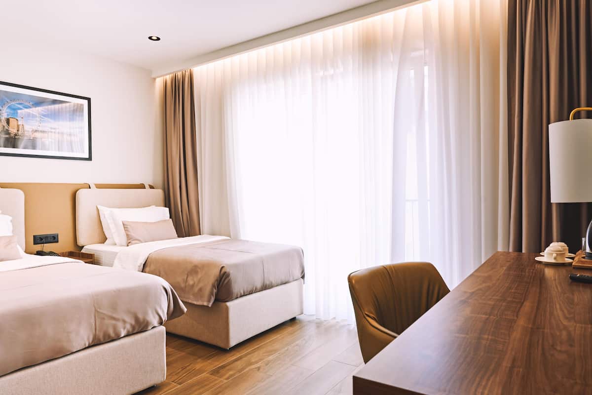 A hotel room with two beds, a wooden dining table, a chair, and a large window with curtains.