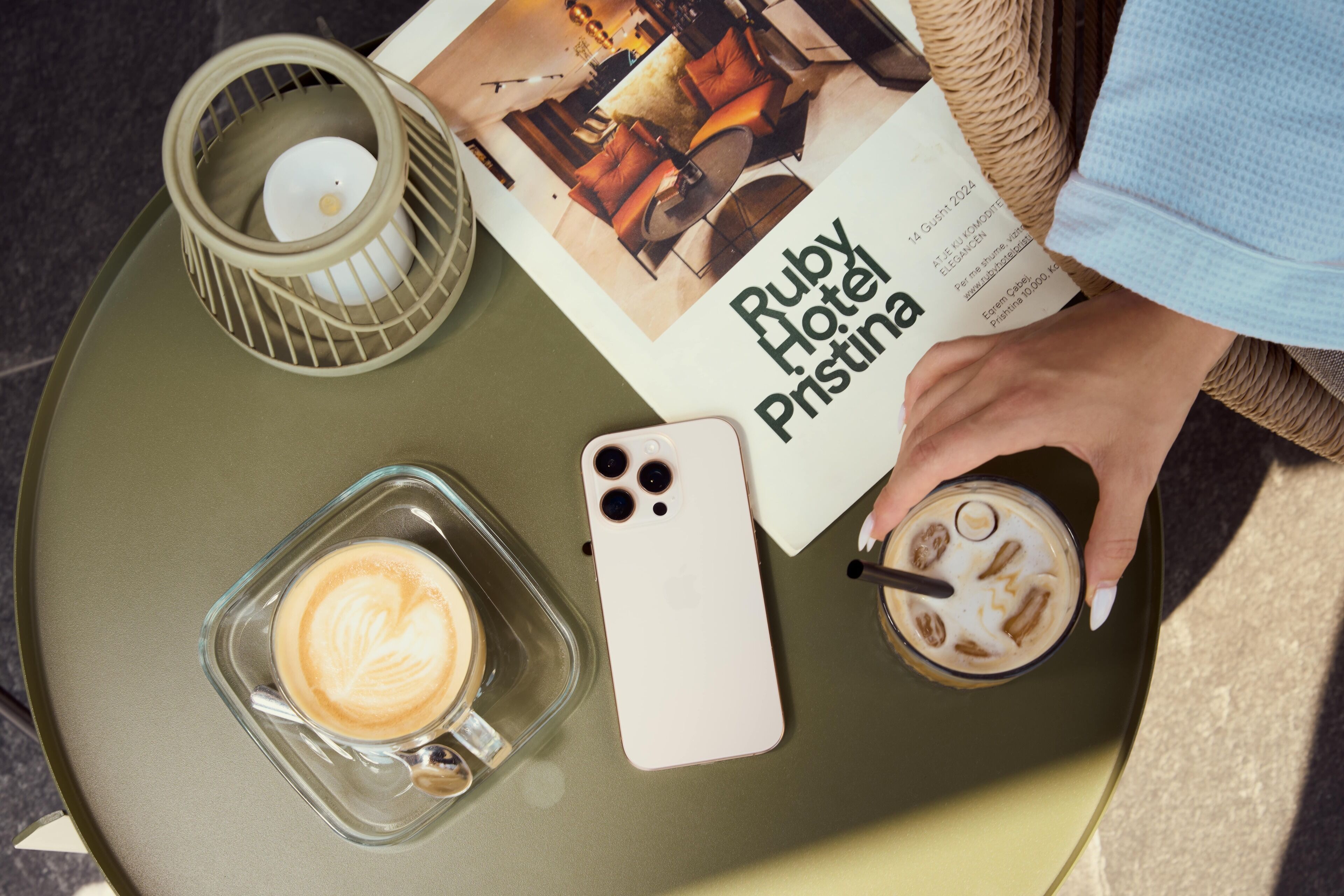 Un tavolo con una tazza da caffè in vetro, uno smartphone, una candela e un menu dell'Hotel Ruby Pristina.