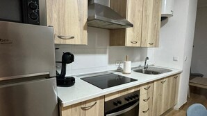 A compact kitchen with a built-in stove, a coffee maker, a sink, and wooden cabinets. - Apartamento Caleta Beach (Cádiz)