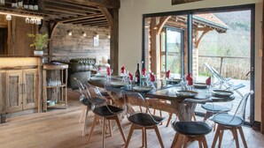 Un comedor rústico con una mesa de madera dispuesta para seis personas, rodeada de sillas transparentes. Un sofá de cuero se encuentra apoyado contra la pared, y se observa un armario de madera a la izquierda.