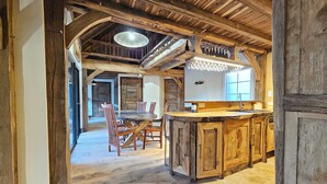 Un interior rústico con una mesa y sillas de madera, una zona de cocina con mostrador de madera, y vigas de madera a la vista en el techo.