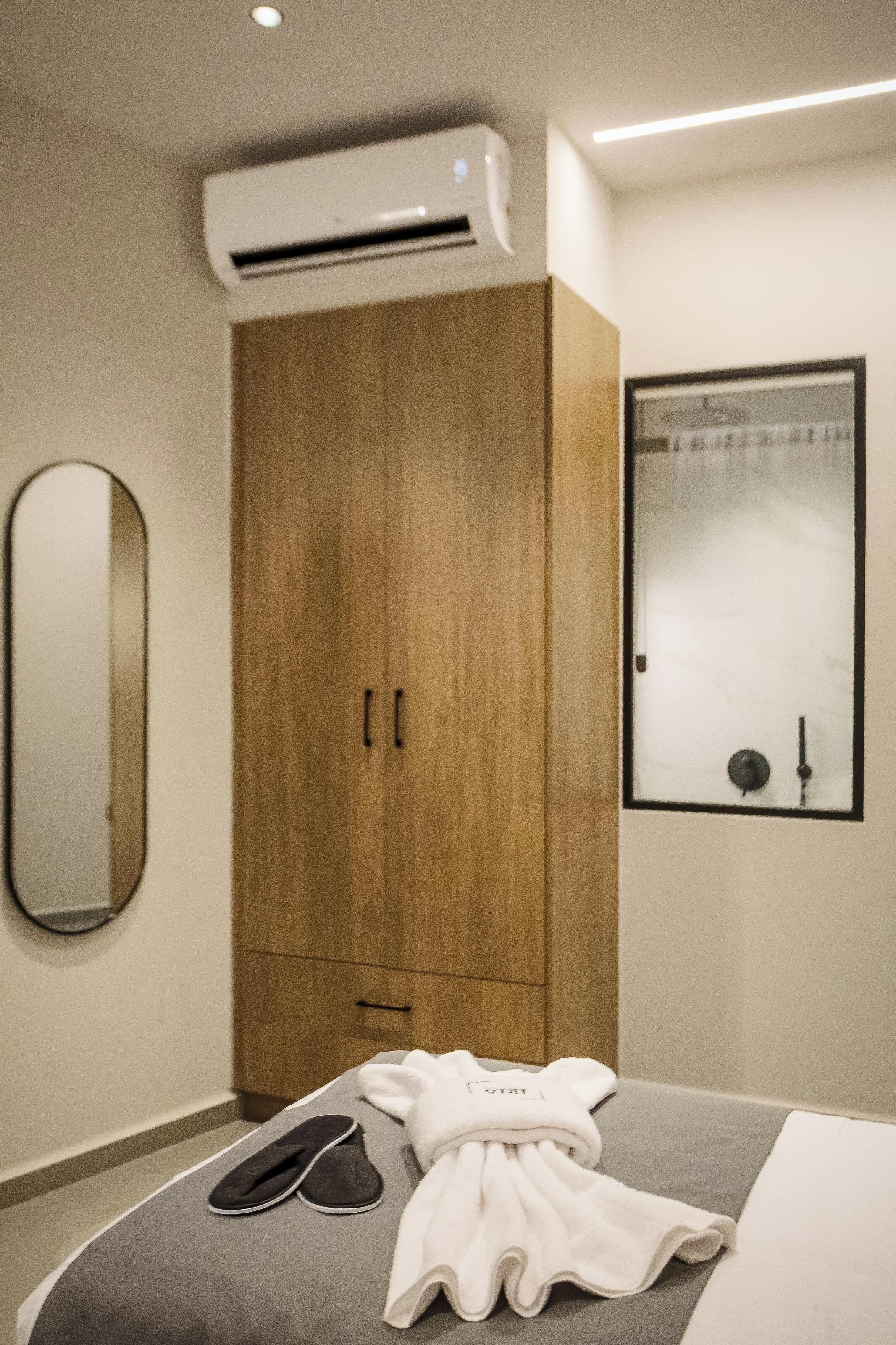 A hotel room with a bed, a wooden wardrobe, a wall-mounted mirror, and an air conditioning unit.