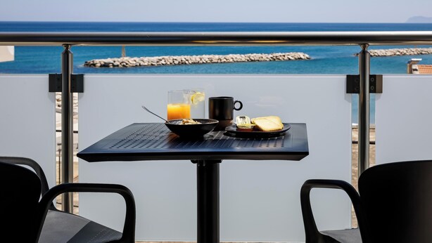 A balcony with a table set for breakfast, overlooking a rocky shoreline and the sea. - Iria Sea View Room (Platanias)