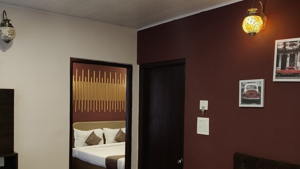 A hotel room with a bed, a small wooden nightstand, a wall-mounted light fixture, and a door leading to another room. - Hotel Route 55 (Darjeeling)