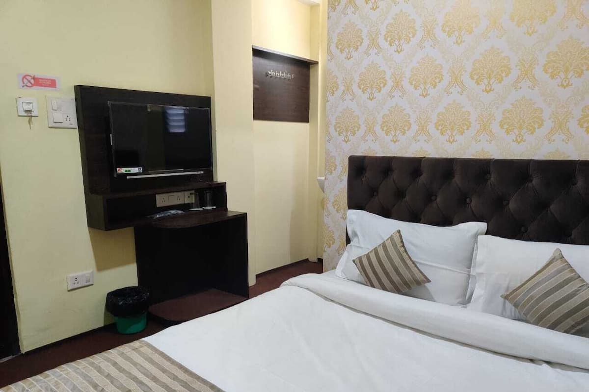 A hotel room with a large bed, a flat-screen TV mounted on the wall, a small table with a lamp, and a trash bin.