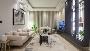 Una sala de estar moderna con un sofá color beige, una mesa de centro y un televisor sobre un mueble bajo. Se observa una estantería negra con objetos decorativos y un ventanal con persianas verticales.