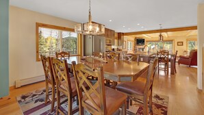 Un comedor amplio con una gran mesa de madera y sillas, una araña y vista a la cocina al fondo.