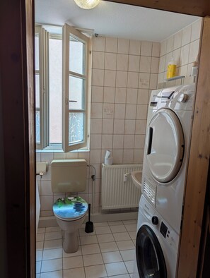 A small, tiled bathroom with a toilet, a washing machine, and a window. - Accommodations in downtown Schwabach for 2 people (Schwabach)