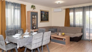 Un salon moderne avec un coin repas, un canapé gris et une table basse. On y trouve une bibliothèque sur laquelle est posée une plante, ainsi qu’un tableau accroché au mur.