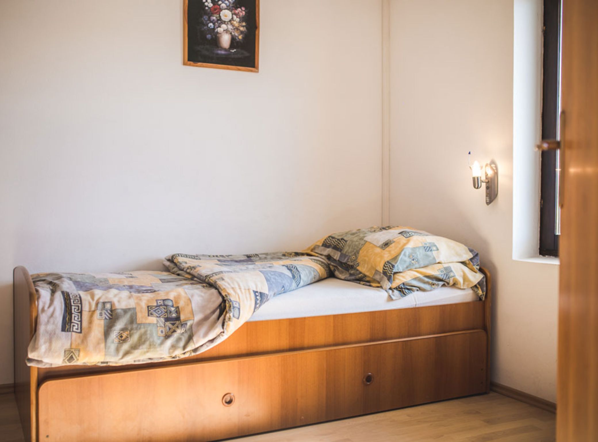 Una cama individual con una colcha y almohadas estampadas, un cabecero de madera, una lámpara de pared y una ventana con vistas al exterior.