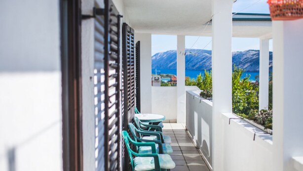 A balcony with a row of green plastic chairs, white walls, and a view of the sea and mountains. - Apartments Jug (Lopar)