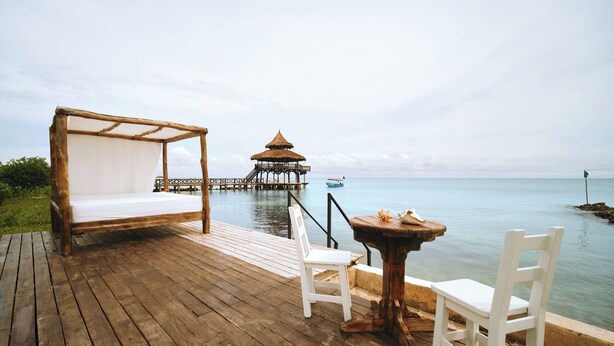 Une terrasse en bois avec un lit, deux chaises et une table, offrant une vue sur une mer calme et une structure distante construite sur pilotis.