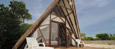 Estructura de madera triangular con puerta de cristal y dos sillas de plástico blanco sobre una terraza de madera.