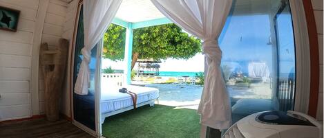 A room with a bed, a window with curtains, a view of the beach, and a small table.