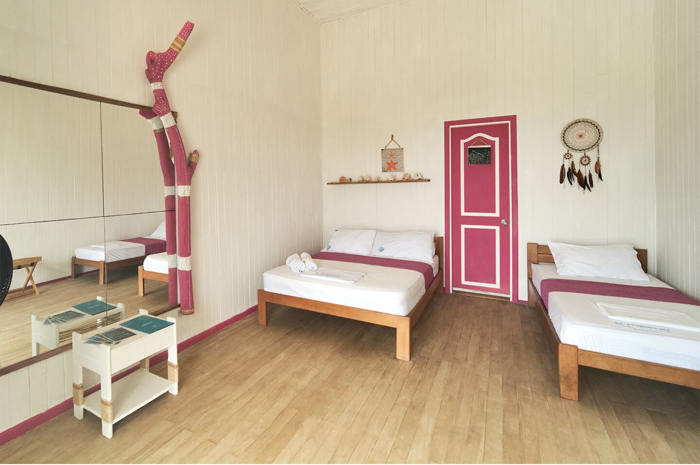Une chambre avec deux lits, un plancher en bois, une porte rose et un attrape-rêves accroché au mur.