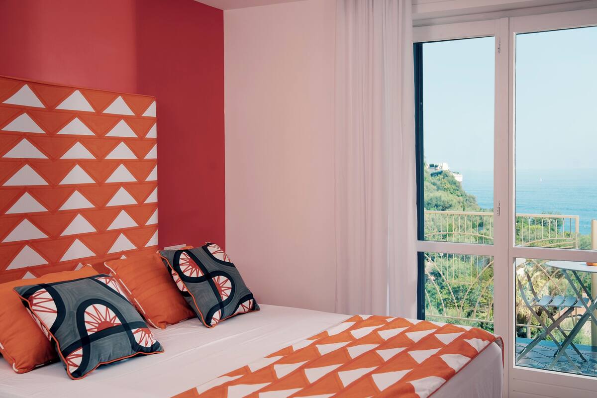 Un dormitorio con una cama grande, almohadas estampadas en naranja y blanco, y vistas al mar a través de amplios ventanales.