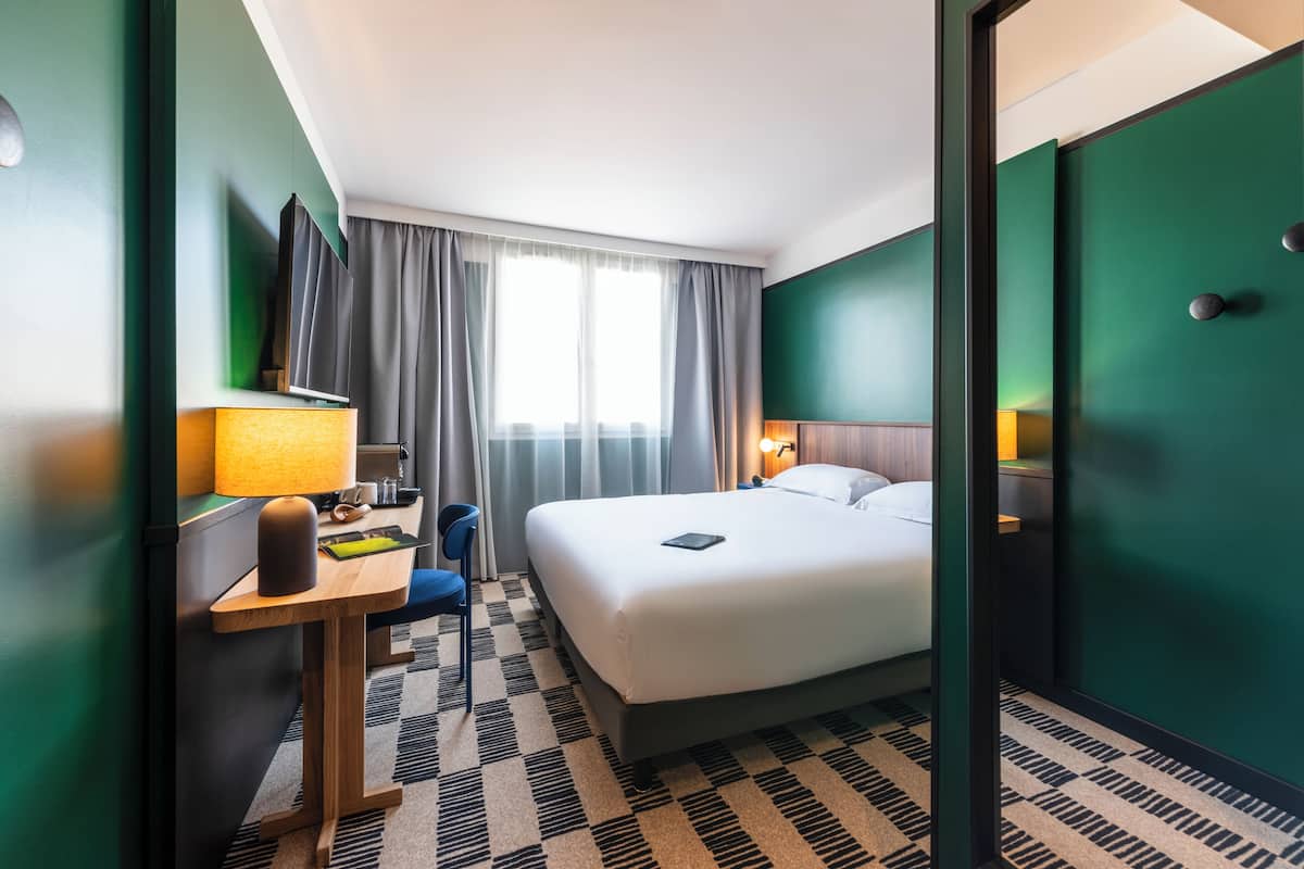 Une chambre d’hôtel avec un lit, un bureau, une chaise, une télévision et une fenêtre avec des rideaux.