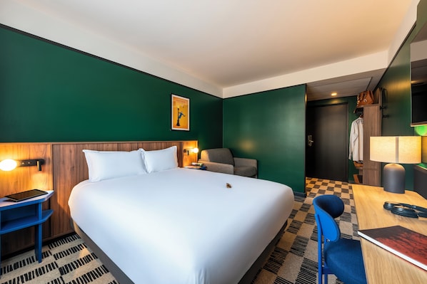 A hotel room with a large bed, a desk with a lamp, a chair, and a small table.
