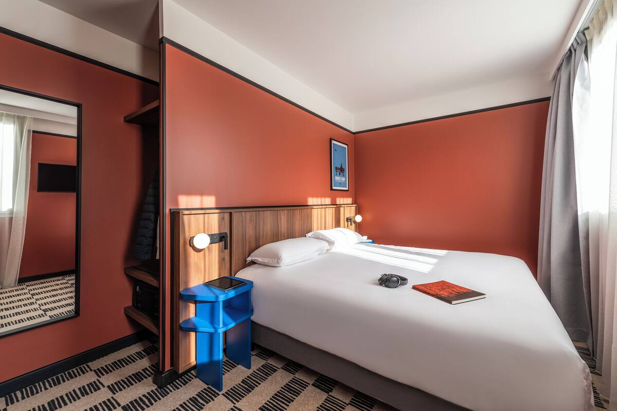 Une chambre d’hôtel avec un lit, une table de chevet, une poubelle bleue, un miroir et un tableau encadré au mur.