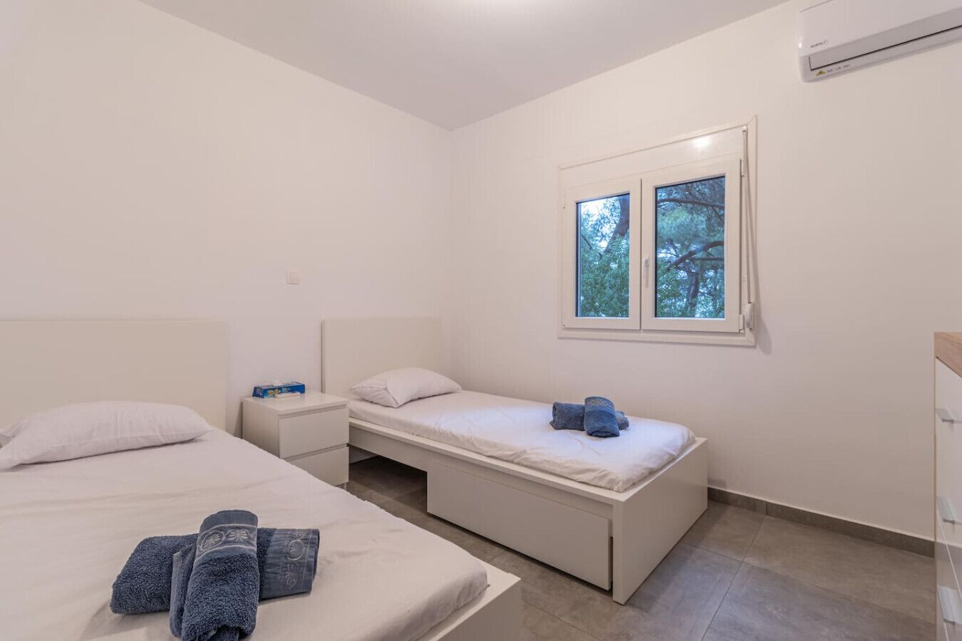 Une chambre à coucher au style minimaliste, avec deux lits, une table de chevet et une fenêtre donnant sur des arbres.