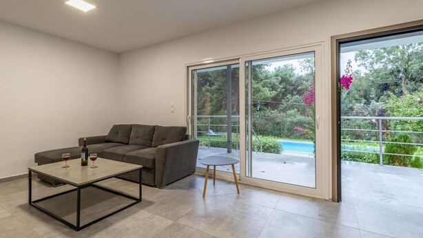 A modern living room with a grey sofa, a coffee table, and a small round table. There is a glass door leading to a balcony with a pool and greenery.