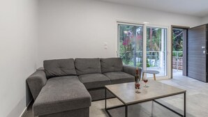 A modern living room with a grey sectional sofa, a glass coffee table, and a view of a balcony with greenery.