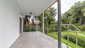 A covered balcony with a metal railing, a concrete floor, and a view of a garden with trees and a house in the background.