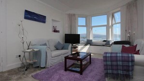 A living room with a sofa, a coffee table, and a TV. There are windows with a view of the sea. - Fairview (Isle of Cumbrae)