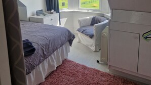 A bedroom with a large bed, a chair, a radiator, and a view of a construction site through a window. - Fairview (Isle of Cumbrae)