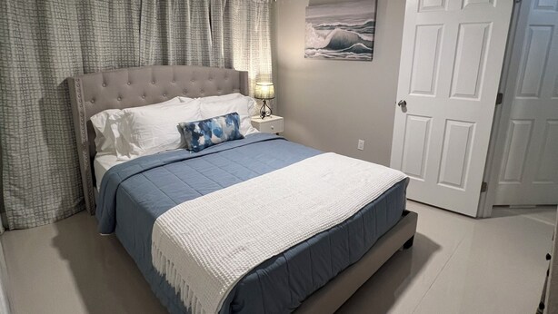 Un dormitorio con cama, ventilador de techo, mesita de noche, lámpara, puerta y un cuadro en la pared.