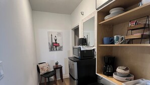 Cocina compacta con refrigerador, microondas y cafetera. Hay un pequeño comedor con una silla y un estante en la pared con vajilla y una planta en maceta.