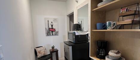 Cocina compacta con refrigerador, microondas y cafetera. Hay un pequeño comedor con una silla y un estante en la pared con vajilla y una planta en maceta.