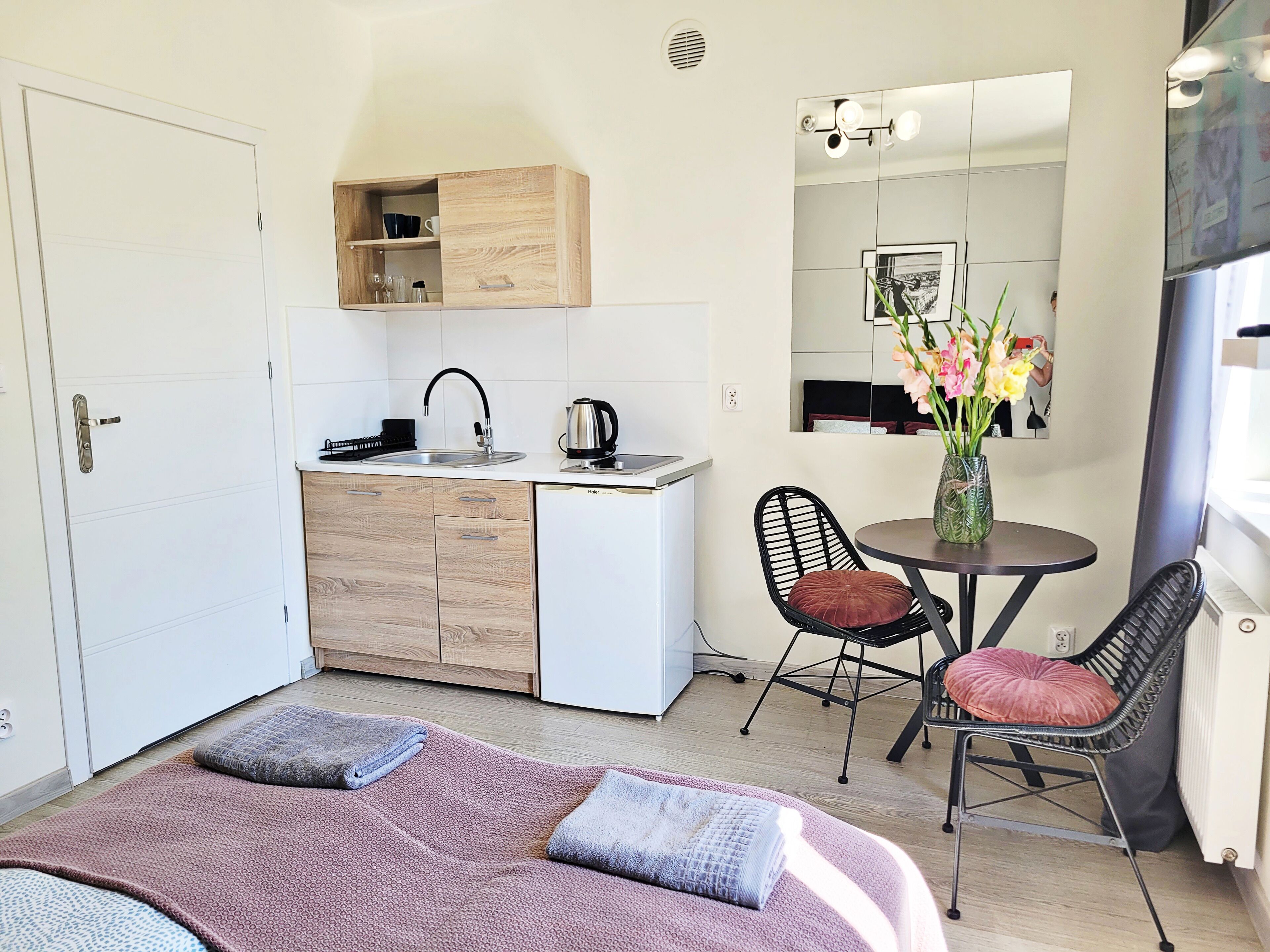 Une chambre d’hôtel compacte comprenant un coin cuisine, un espace repas avec deux chaises, un lit recouvert d’une couverture à motifs et un miroir qui reflète la pièce.