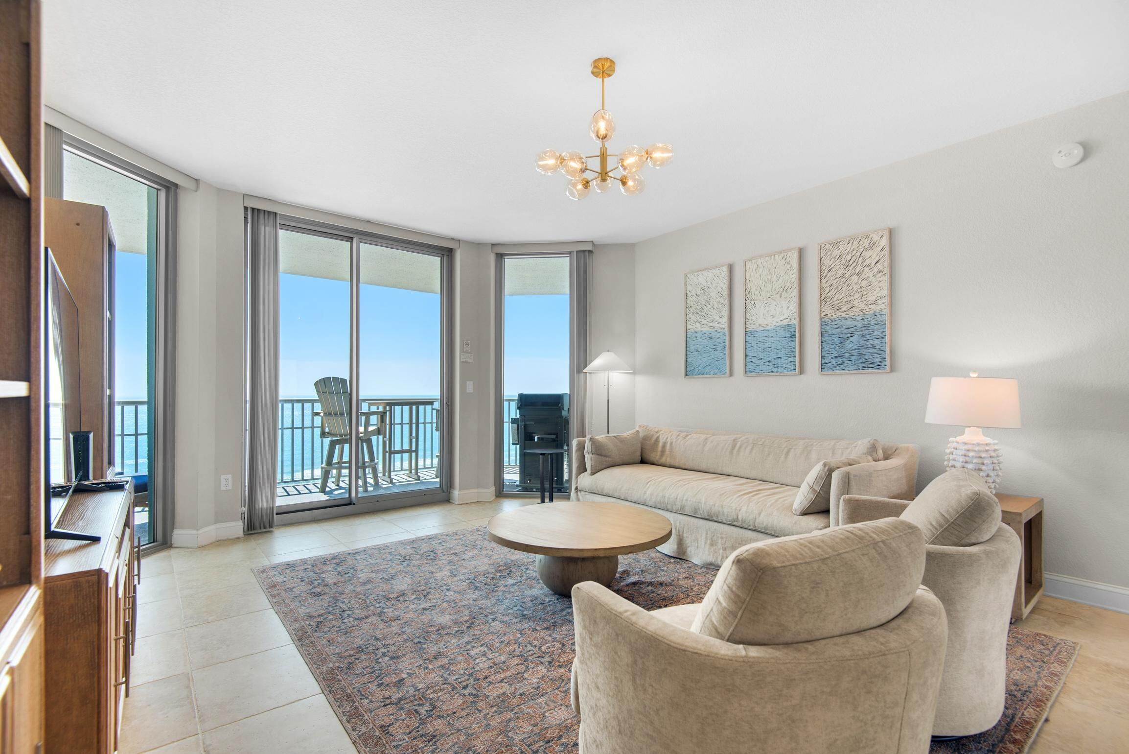 A spacious living room with a sofa, armchairs, a coffee table, and a chandelier. There is a balcony with a view of the ocean.