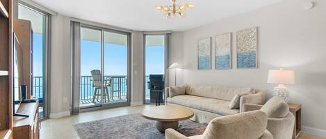 A spacious living room with a sofa, armchairs, a coffee table, and a chandelier. There is a balcony with a view of the ocean.