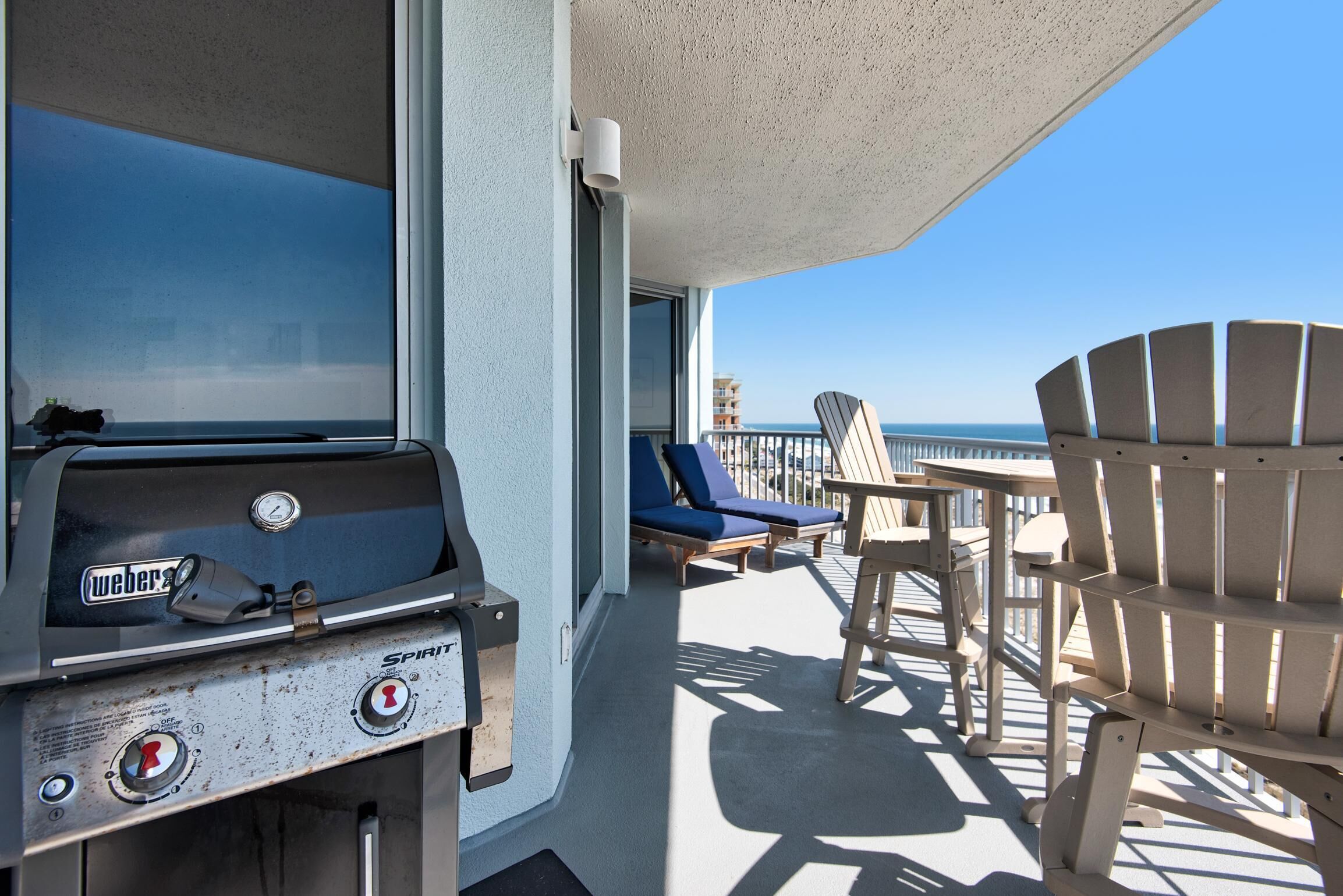 A balcony with a Weber Spirit grill, blue lounge chairs, and a view of the ocean.