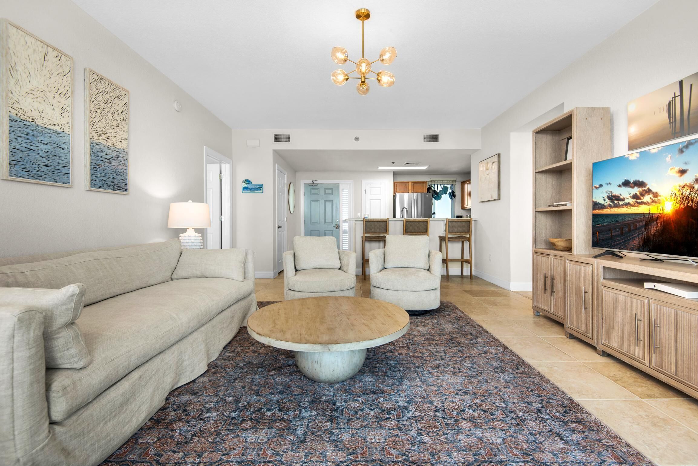 Un salon spacieux avec un canapé beige, deux fauteuils, une table basse, un téléviseur et des œuvres d’art accrochées aux murs.