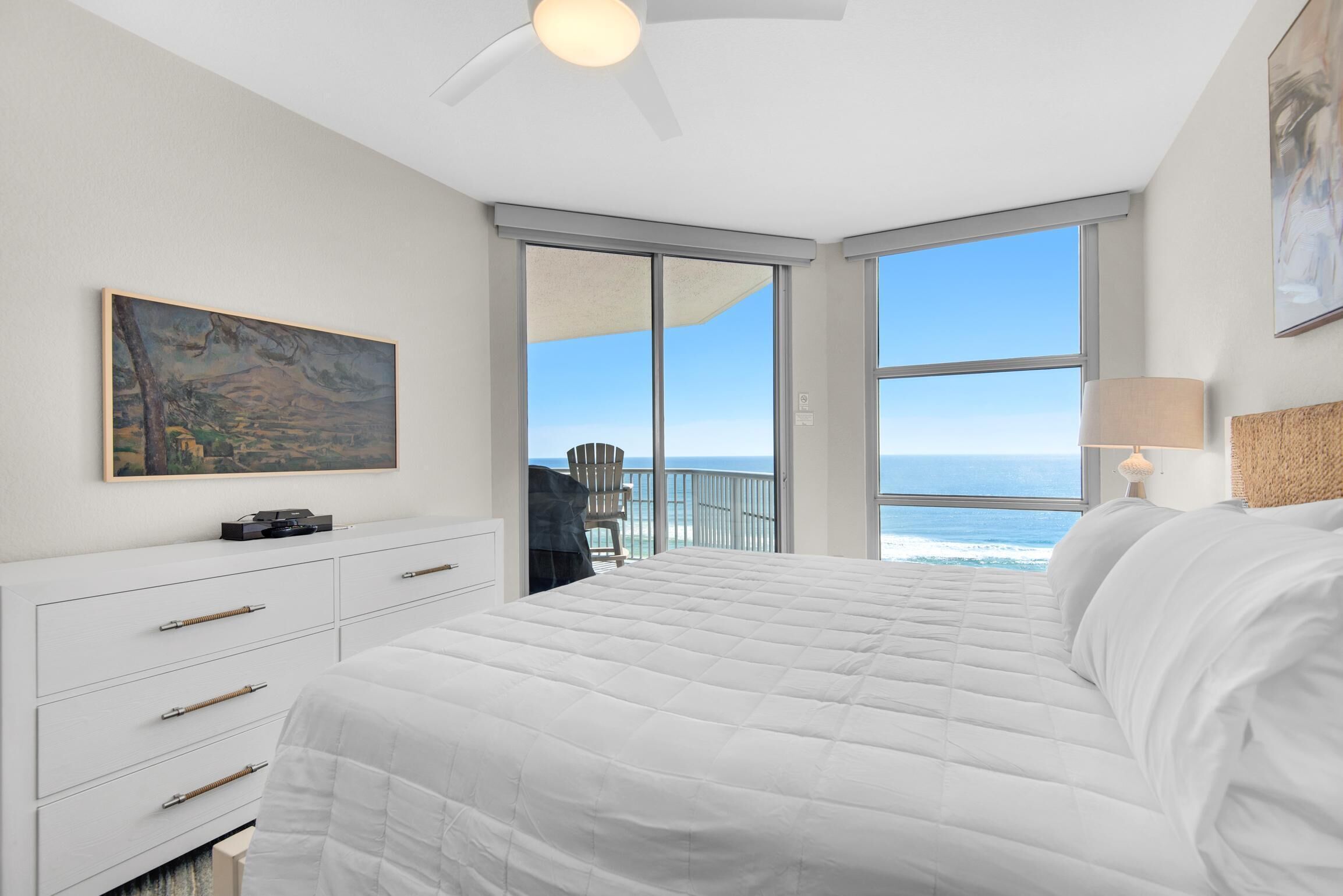A bedroom with a large bed, a dresser, a painting, a chair, and a view of the ocean through a sliding door.