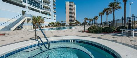 A swimming pool area with a clear blue pool, steps leading into the pool, a palm tree-lined walkway, and a high-rise building in the background.