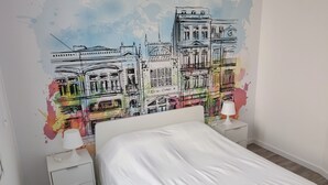 Ein Schlafzimmer mit einem weißen Bett, zwei Nachttischen mit Lampen und einem farbenfrohen Stadtbild-Wandgemälde.