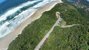 An aerial view of a coastal area with a beach, a road winding through a forested hillside, and a clear blue sky. - Pousada Recanto do Rosa (Imbituba)