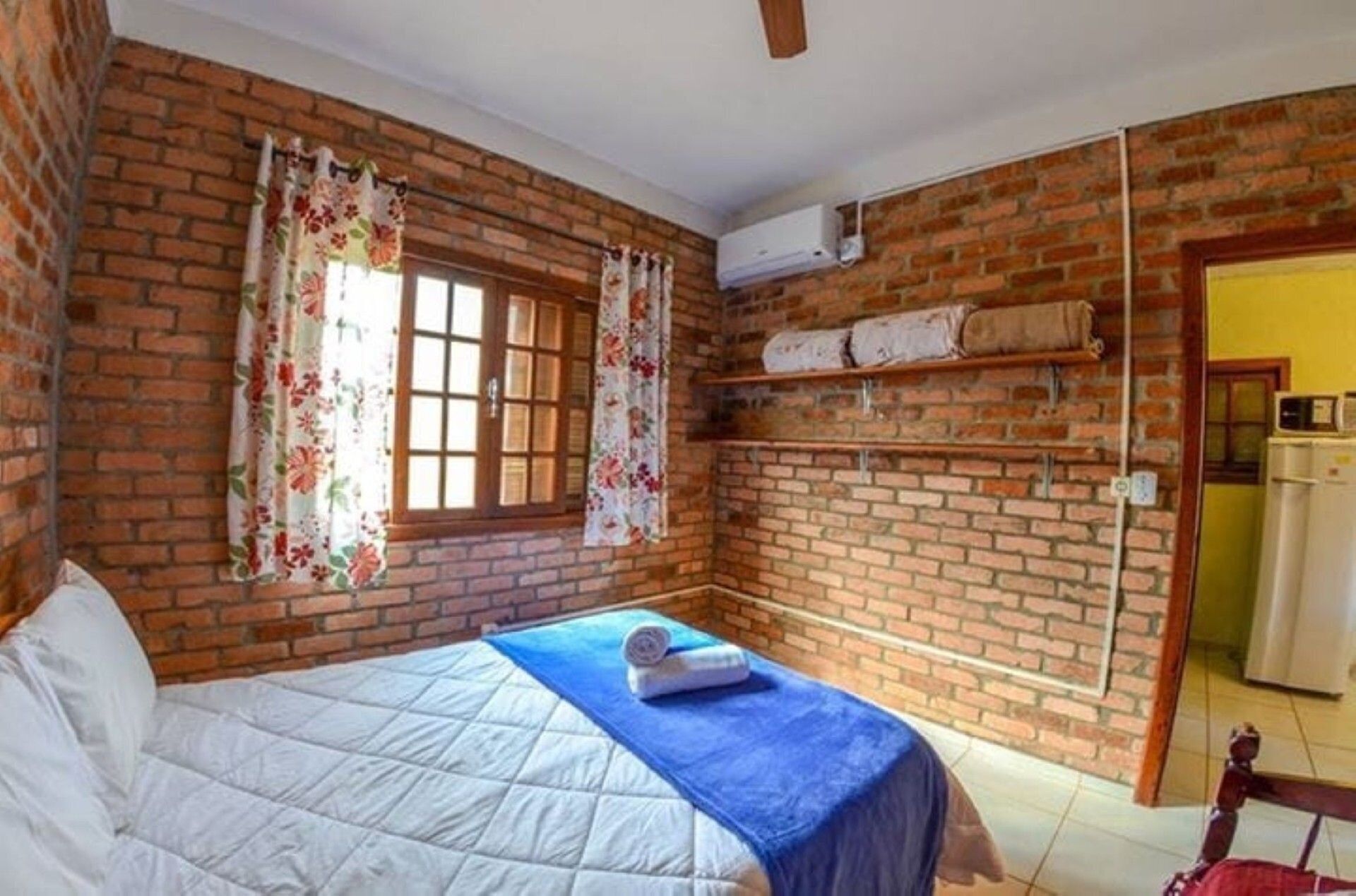 A room with a brick wall, a bed with a blue blanket, a window with floral curtains, and a built-in shelf with towels.