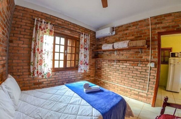 A room with a brick wall, a bed with a blue blanket, a window with floral curtains, and a built-in shelf with towels. - Pousada Recanto do Rosa (Imbituba)