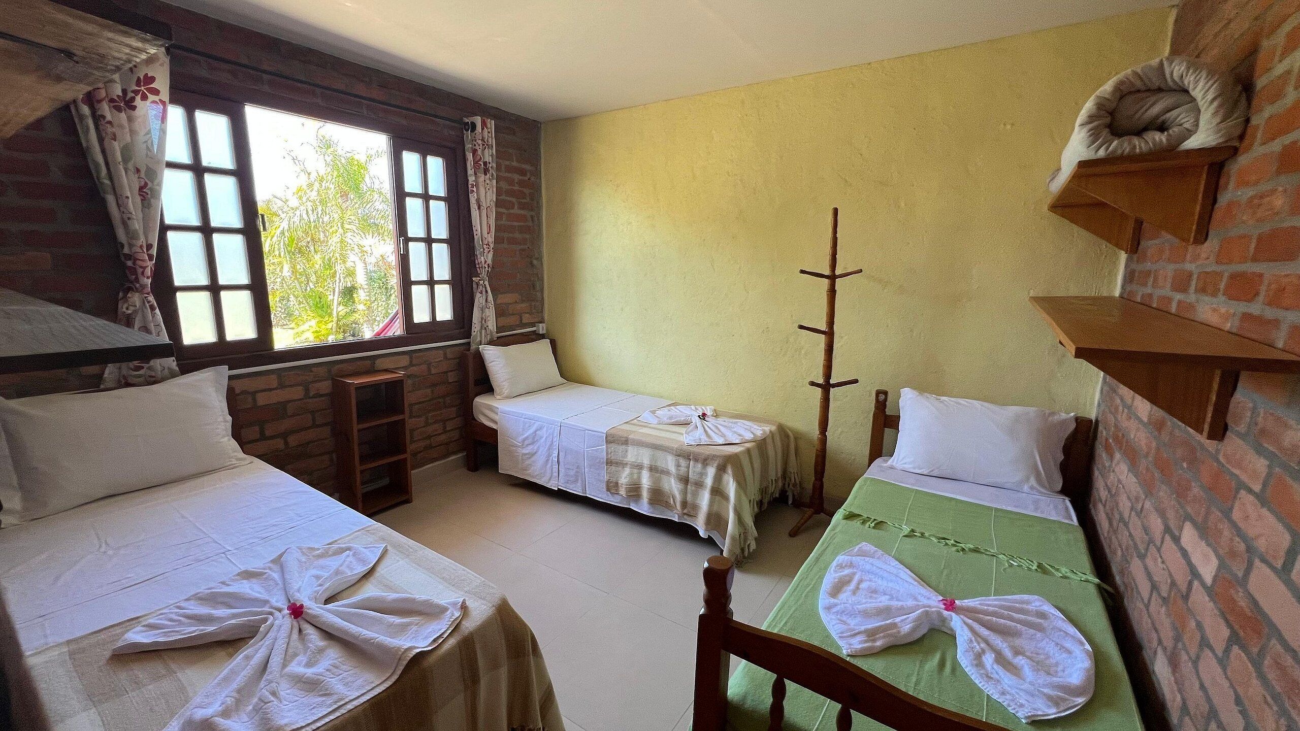 A room with two single beds, each with a towel on the bed. There is a window with curtains, a brick wall, and a wooden shelf.