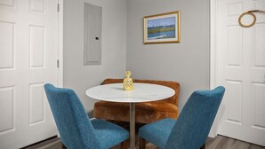 Un coin repas avec une table ronde, deux chaises bleues, un ananas en guise de décoration centrale et un tableau encadré au mur.