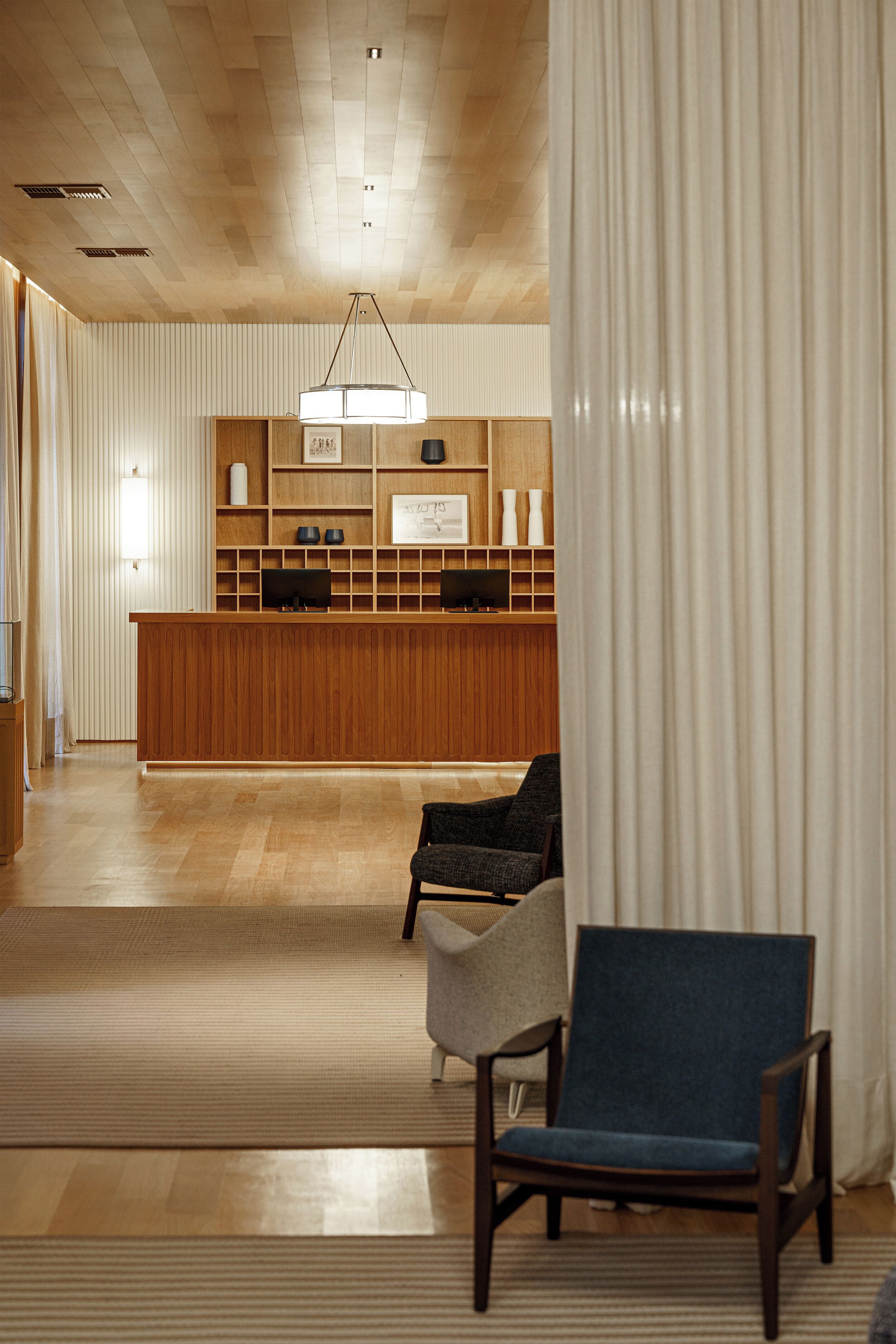A reception area with a wooden desk, two chairs, a hanging light fixture, and a large window with curtains.