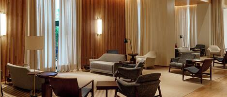 A hotel room with a large bed, multiple seating areas, and a view of greenery through the curtains.