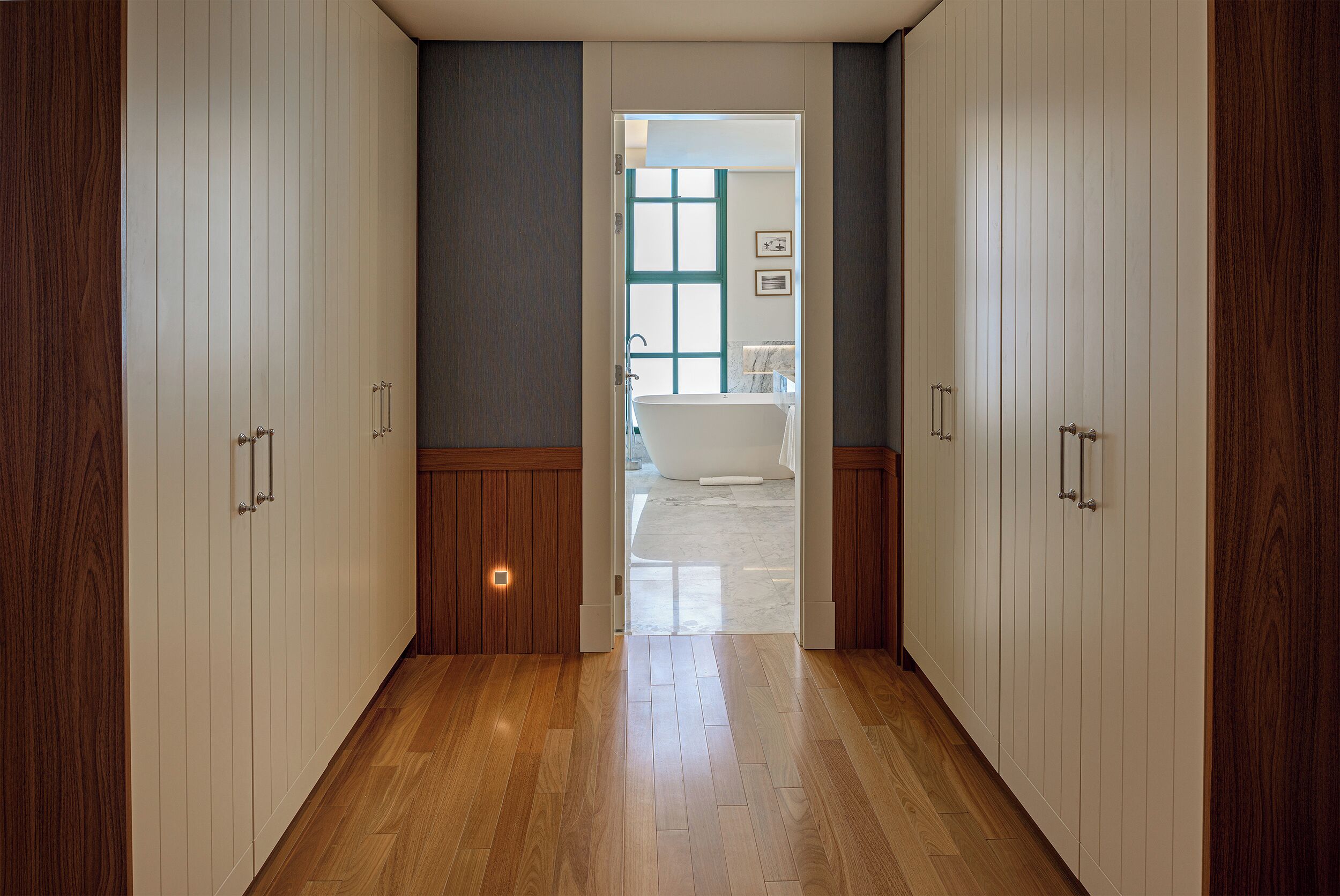 A hallway with wooden flooring leading to a bathroom with a bathtub and a window.