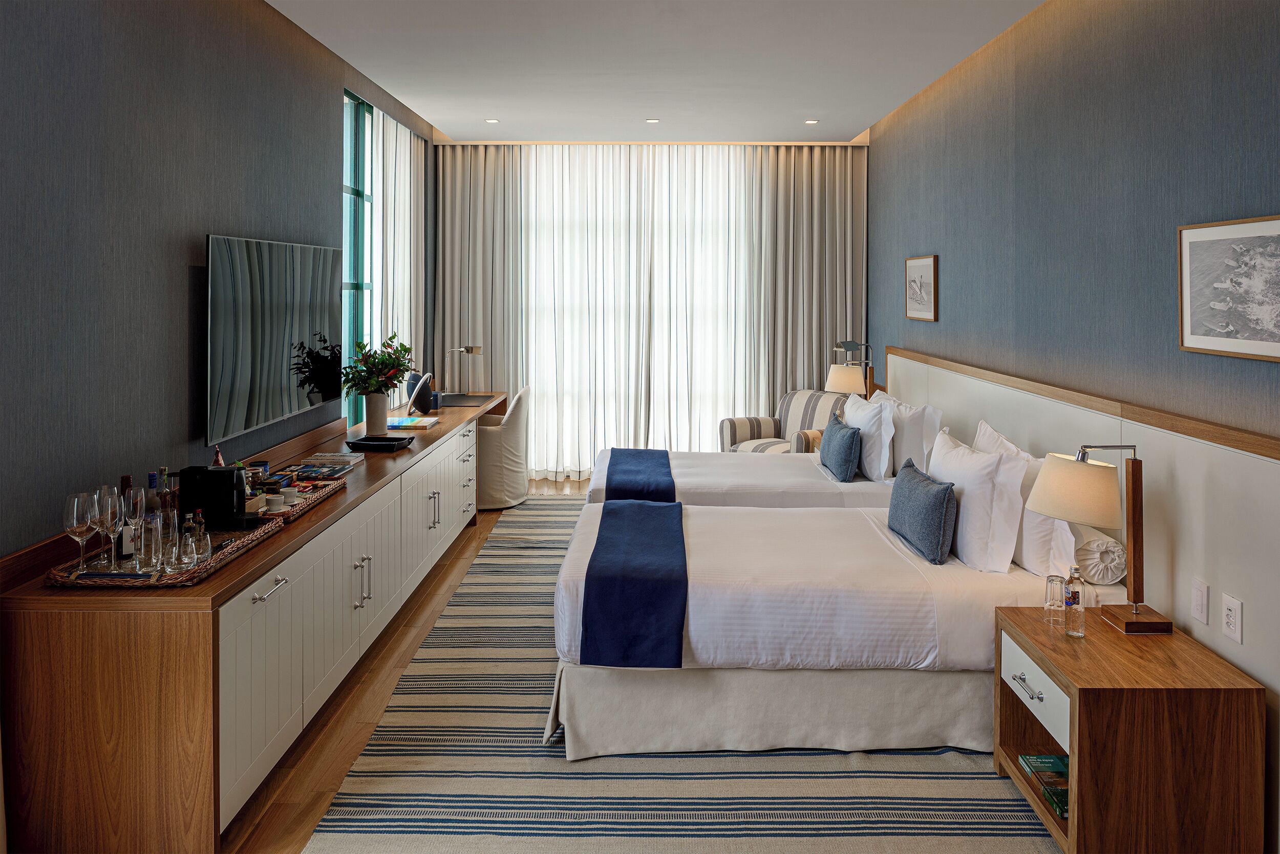 Una habitación de hotel con una cama grande, una cama más pequeña, un escritorio con computadora, un televisor y una ventana con cortinas.