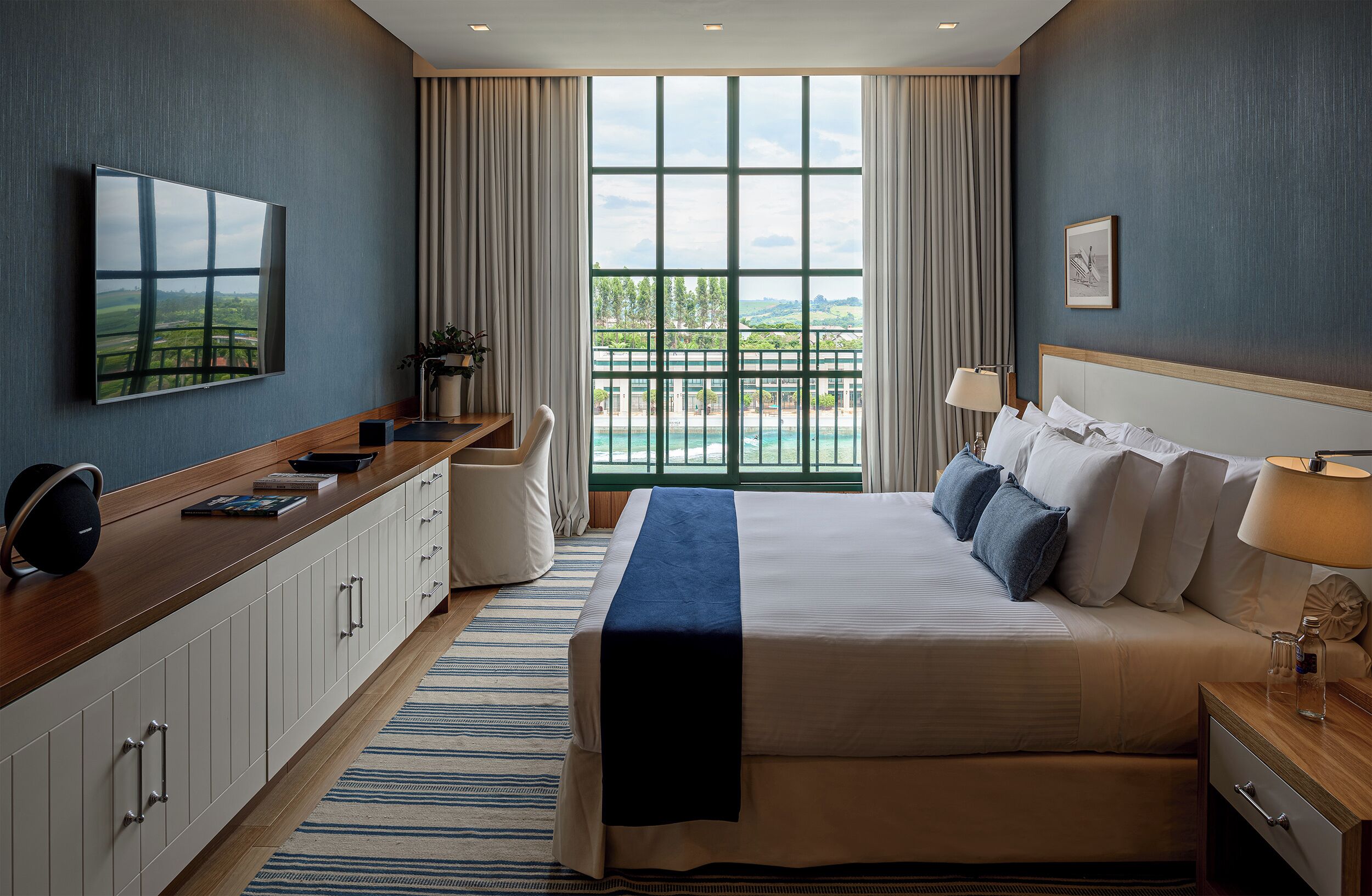 A hotel room with a large bed, a TV mounted on the wall, a desk with a computer, and a view of a pool through the window.